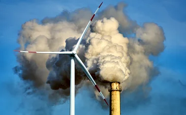 Wind turbine with a smoking chimney behind it 