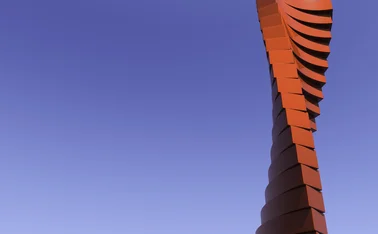 A tall, modern red sculpture with a twisting, stacked design rises against a clear blue sky, featuring smooth, geometric layers that create a dynamic spiral effect, standing out vividly due to its bold red colour and minimalist backdrop.