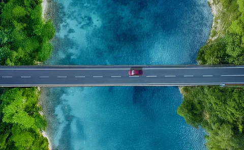 Aerial view of a red car driving across a straight bridge over crystal-clear turquoise water, surrounded by dense green forest on both sides.