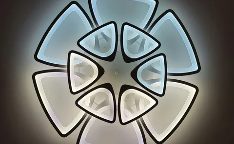 Modern LED light graphic featuring a dramatic geometric floral design. The fixture is composed of multiple tiers of illuminated triangles with black borders, creating a stylized flower pattern against a dark background.