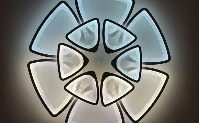 Modern LED light graphic featuring a dramatic geometric floral design. The fixture is composed of multiple tiers of illuminated triangles with black borders, creating a stylized flower pattern against a dark background.