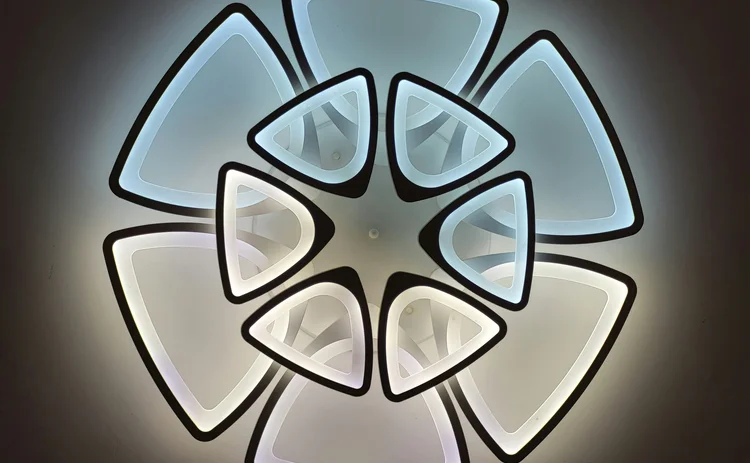 Modern LED light graphic featuring a dramatic geometric floral design. The fixture is composed of multiple tiers of illuminated triangles with black borders, creating a stylized flower pattern against a dark background.