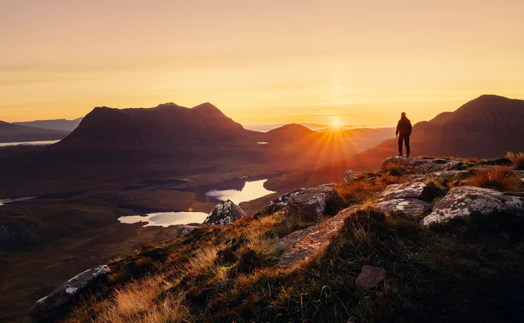 Hiker standing on a rocky mountain ridge at sunrise, overlooking a scenic valley with reflective lakes and rugged peaks under a golden sky, symbolizing adventure, exploration, and natural beauty