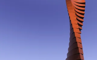 A tall, modern red sculpture with a twisting, stacked design rises against a clear blue sky, featuring smooth, geometric layers that create a dynamic spiral effect, standing out vividly due to its bold red colour and minimalist backdrop.